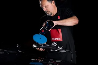 Man polishing a black car with a blue buffer in a dark setting.