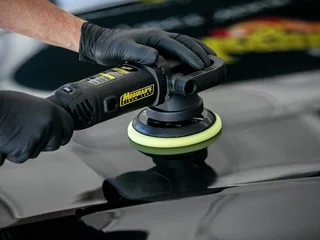Gloved hand polishing a black car hood with a rotary buffer.
