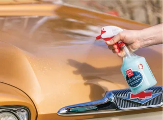 Hand spraying cleaner on the hood of a gold car near the windshield.