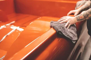 Tattooed person wiping an orange car hood with a gray cloth.