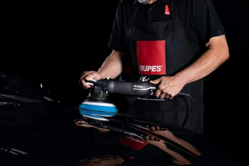 Man polishing a black car with a power buffer against a dark background