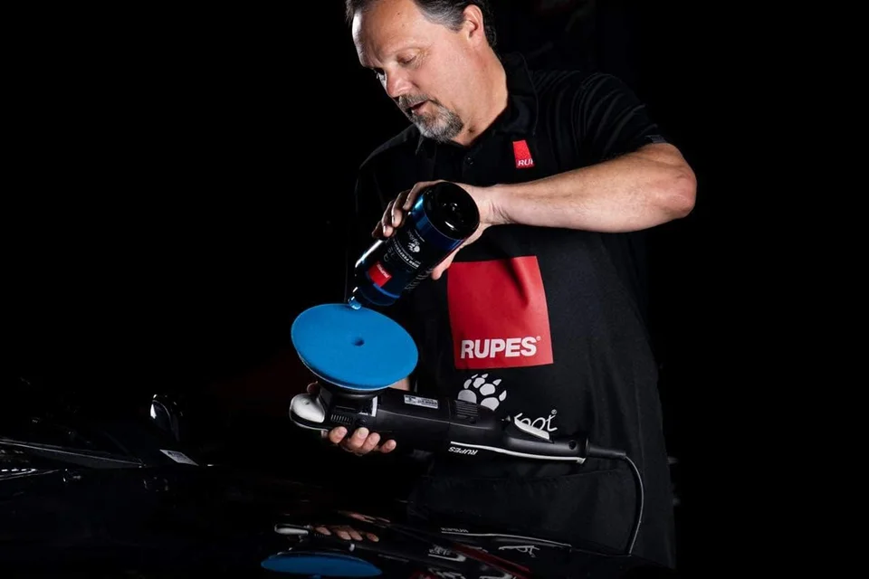 Man polishing a black car with a blue buffer in a dark setting.
