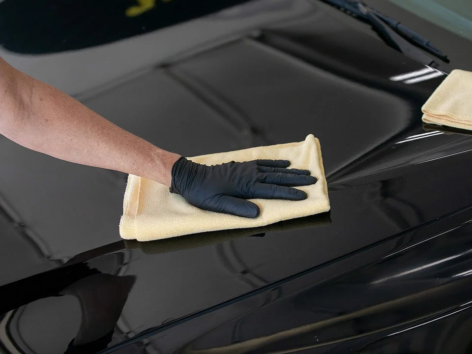 Person in black glove wiping a shiny black car hood with a yellow cloth